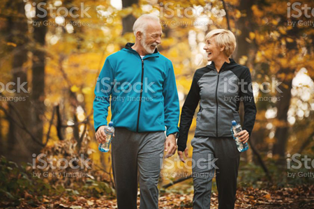 Um homem e uma mulher da melhor idade caminham em um parque, se entreolham e sorriem.
